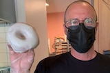 A man in a black face mask holds a white-iced doughnut with a gloved hand.