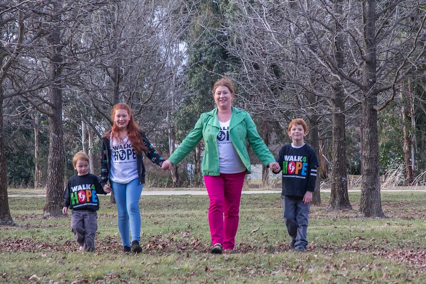 A woman with redhead holding hands with three children with red hair walking through an avenue of trees
