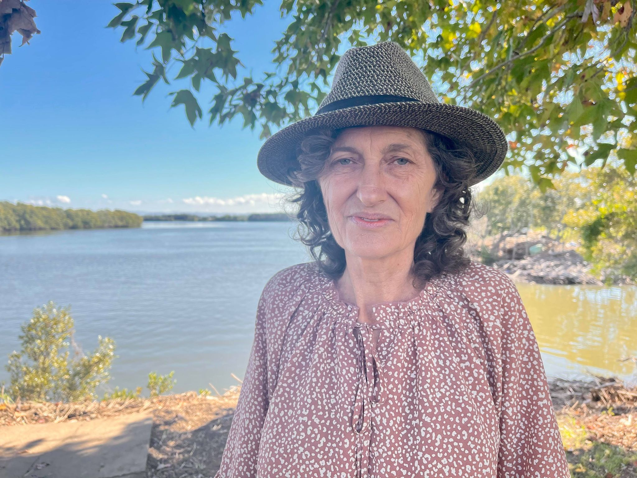 Woman in a pink shirt and grey hat stands under a tree in front of a river.