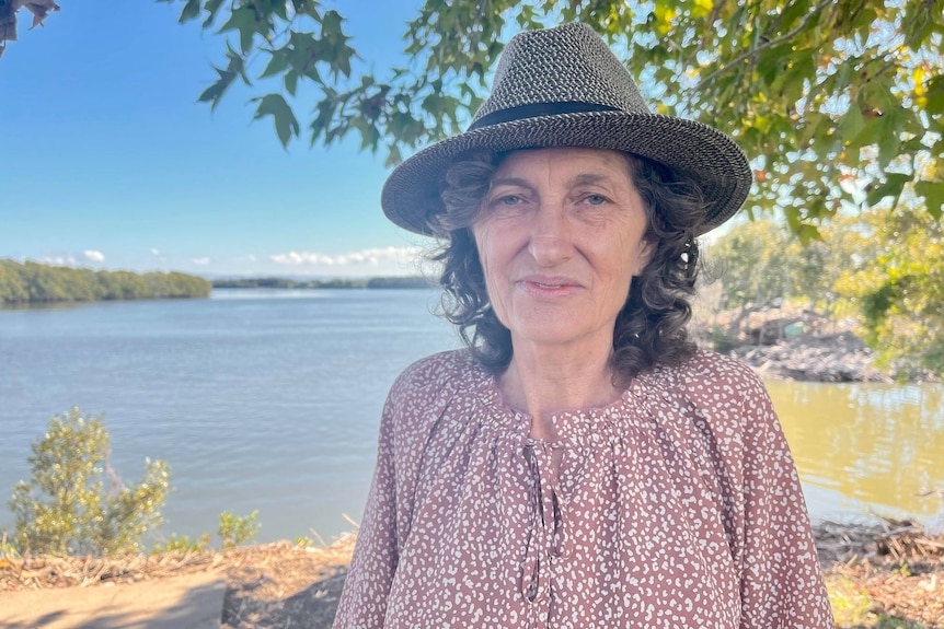 Woman in a pink shirt and grey hat stands under a tree in front of a river.