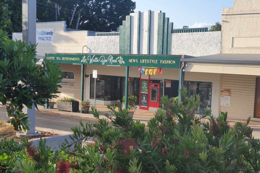 A beige and green building for a newsagency. There are plants in the foreground of the photo. The newsagency has a red ATM.