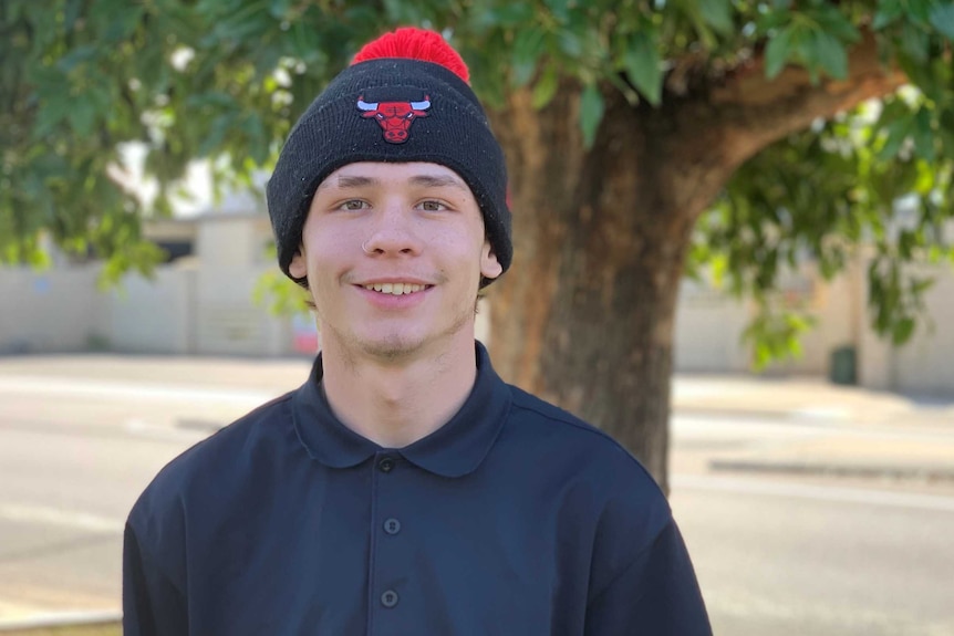 A young man with a beanie on standing in front of a tree with a road in the background.