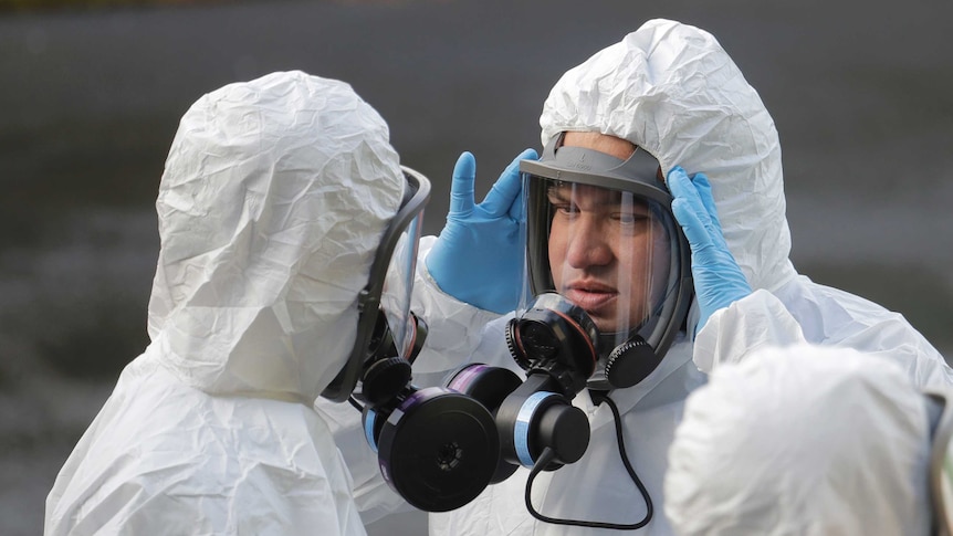 A man wearing a clear respirator, a white protective suit and blue gloves adjusts his mask.