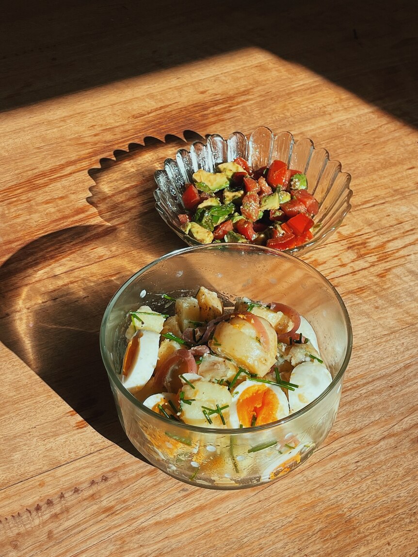 A glass bowl of nicoise salad below another bowl of avocado tomato salad on a timber dining table, drenched in golden light.