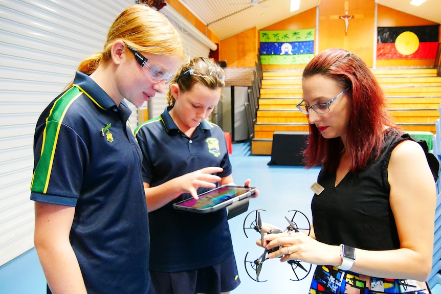 A female teacher holds a drone, and instructs two female primary students how to use it, as they hold an ipad.