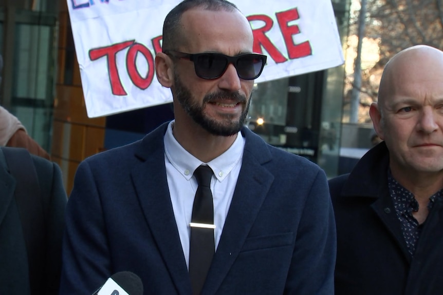 Three men in suits standing in front of a protester's sign