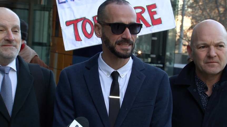 Three men in suits standing in front of a protester's sign