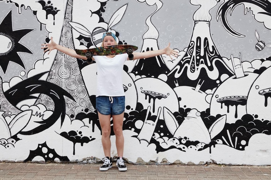 A young girl with a skateboard stands with her arms spread wide, in front of a wall covered in a black-and-white mural.