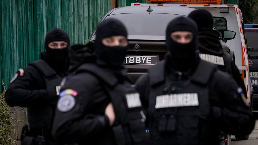 Romanian gendarmes in black clothing and masks are backdropped by a UK registered luxury vehicle.