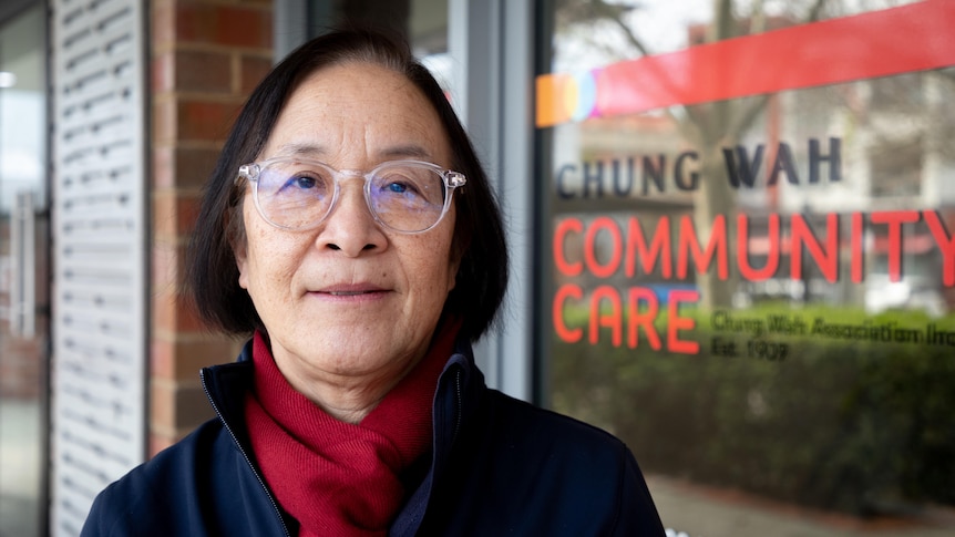 A woman with glasses stands in front of a building with a sign reading Chung Wah.