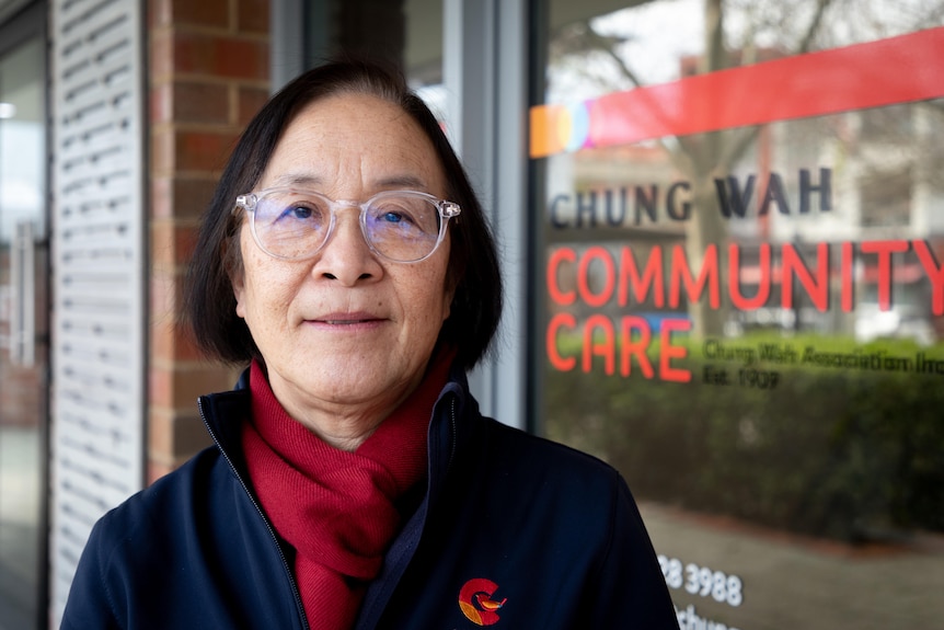 A woman with glasses stands in front of a building with a sign reading Chung Wah.