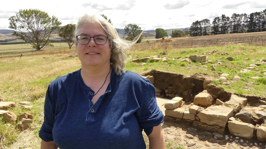 A grey-haired woman in glasses and blue top stands in a paddock in front of an archaeological dig.
