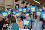 A group of mainly elderly people hold of 'VOTE NO' placards in a room. There are hundreds of people.