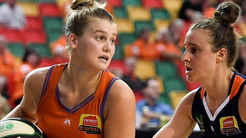 A WNBL basketballer holds the ball and looks around for a teammate while an opponent guards her.