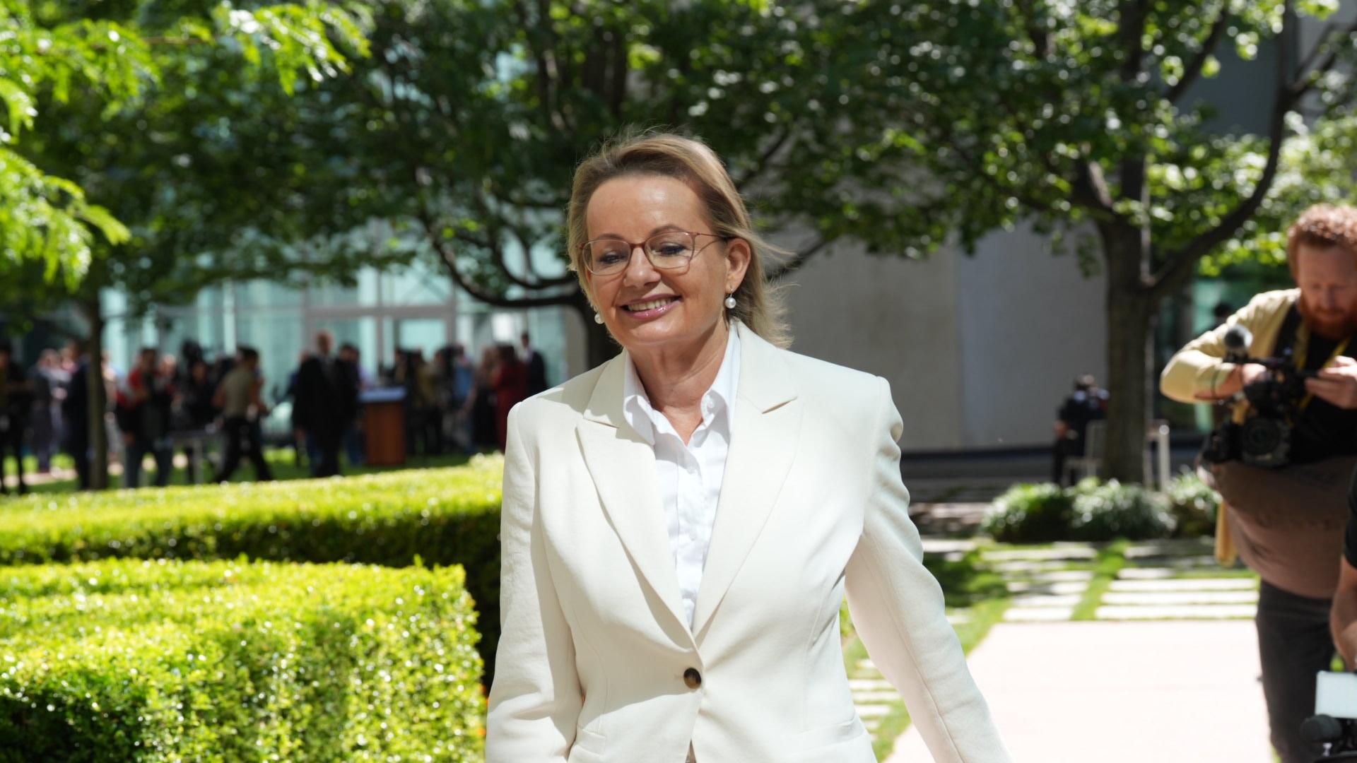 Una mujer rubia sonriente con un traje ligero camina por un sendero bordeado de setos.