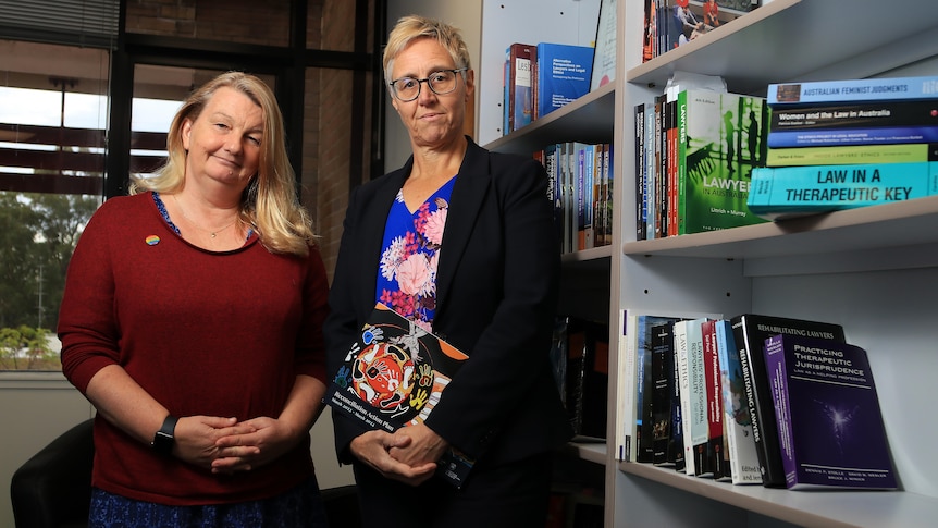 Nancy Huggett wears a maroon jumper and Trish Mundy wears a black blazer with floral shirt, holding a law textbook in an office.