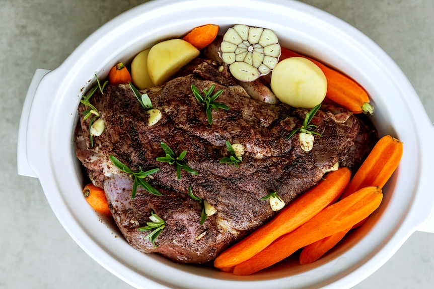 Cooked lamb leg in a slow cooker surrounded by carrots and potatoes, an easy weeknight family meal.