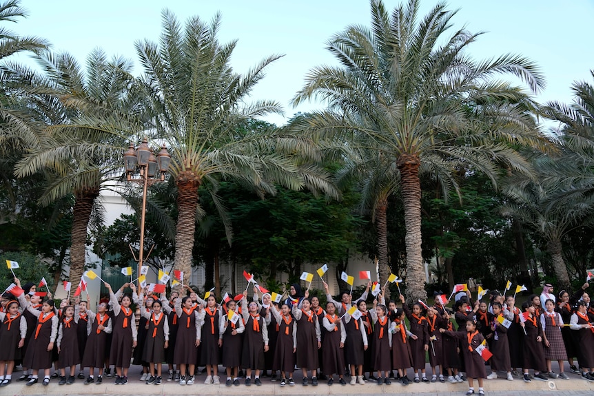 Children in school uniform wave flags while standing under large palm trees. 
