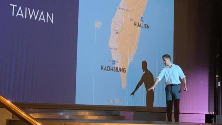 A man on a stage in front of a projection of a map of Taiwan