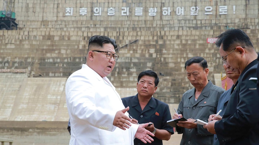 Kim Jong-un stands with mouth open and arms outstretched by his hips, looking at four men standing close taking notes, outdoors