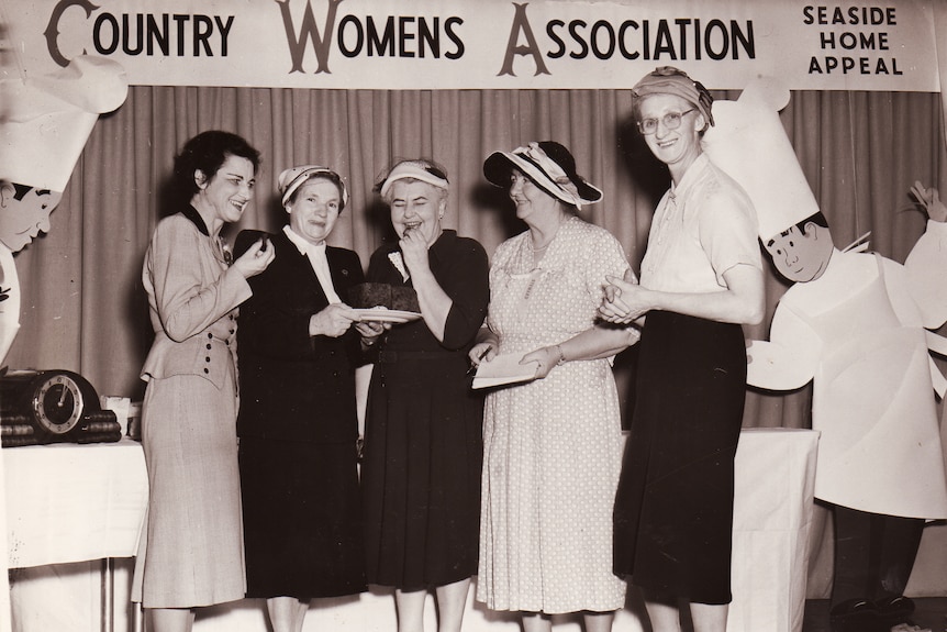 A black and white photograph of five smiling women standing in a hall with a Country Women's Association banner behind.