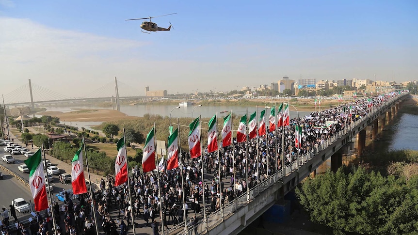 A wide shot shows hundreds of protestors walking down an Iranian street