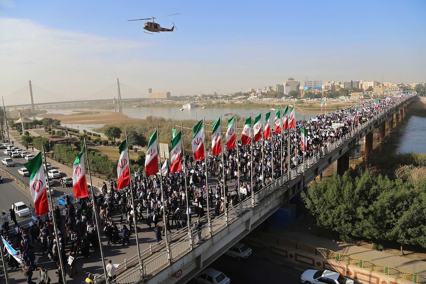 A wide shot shows hundreds of protestors walking down an Iranian street