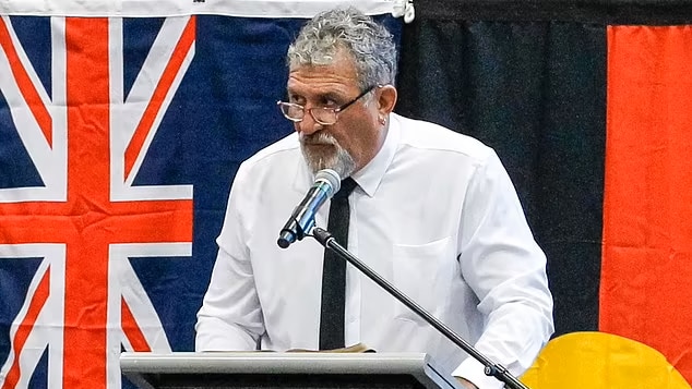 A man in a white shirt, black tie, wearing glasses stands in front of Australian and Aboriginal flags, speaking in a microphone.