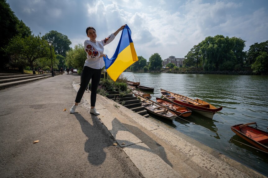 A woman waves a yellow and blue Ukrainian flag while standing next to a canal with row boats tied up near the edge