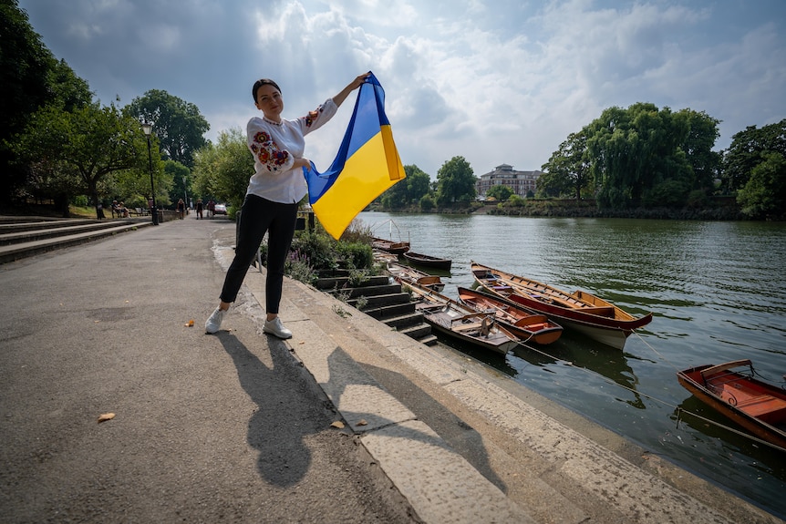 Женщина машет желто-синим украинским флагом, стоя рядом с каналом с привязанными к краю гребными лодками.