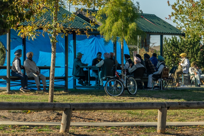 A group of people, many dressed in turbans, sit under an outdoor shelter. A blue tarpaulin hangs from the roof on one side. 