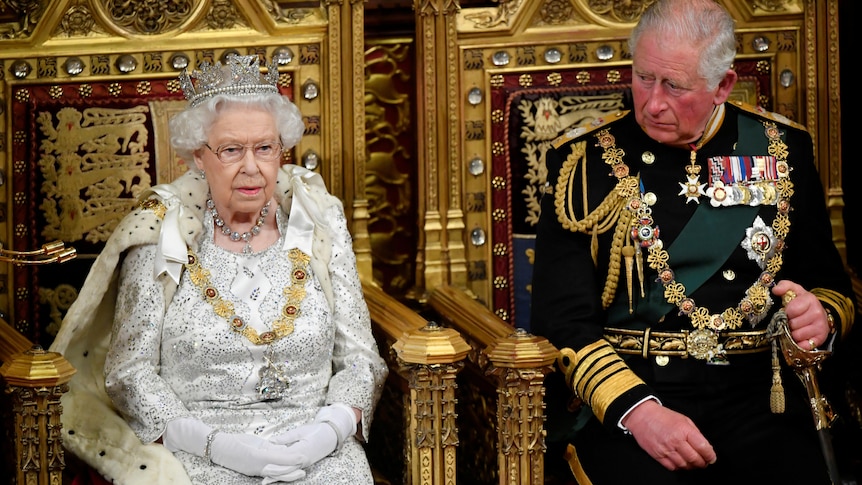Queen Elizabeth in a tiara and white dress sits in a golden throne while Prince Charles sits next to her
