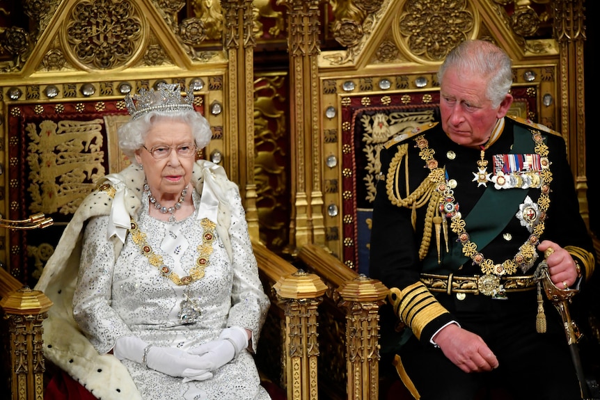 Queen Elizabeth in a tiara and white dress sits in a golden throne while Prince Charles sits next to her