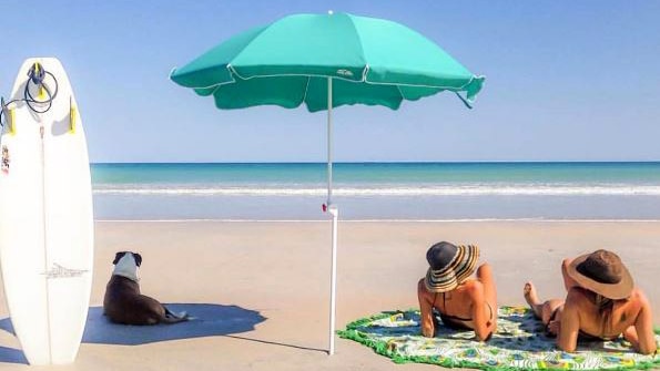 Two women lie on a mat at Cable Beach with a dog, an umbrella, and a surfboard nearby.