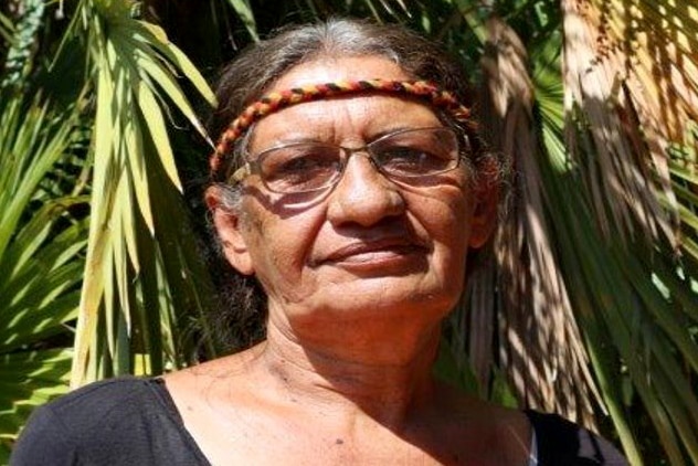 Bonnie Edwards wears a red, yellow and black headband and smiles slightly, standing in front of a palm tree.