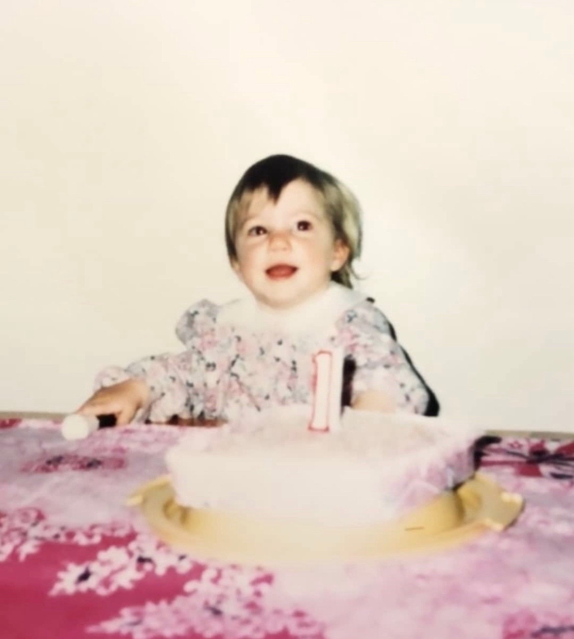 An old coloured photo of a child, smiling, sitting in front of birthday cake, 1 candle on it.