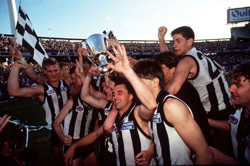 Collingwood captain holds premiership cup above his head as players celebrate with black and white checked flag in background. 