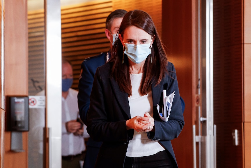 Jacinda Ardern in a face mask, clasping her hands as she walks through doors 