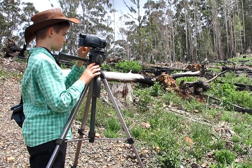12 year old boy standing being video camera shooting toward a patch of clear-felled forest