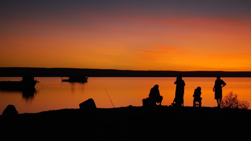 Four people silhouetted against a setting sun and orange sky fish in a bay at Wyndham with two boats on the water.