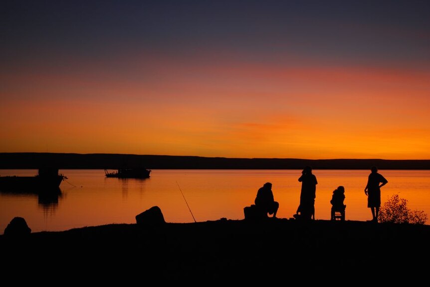 Four people silhouetted against a setting sun and orange sky fish in a bay at Wyndham with two boats on the water.