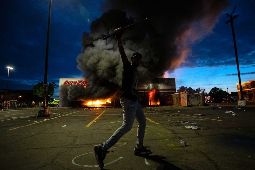 A man poses for a photo in the parking lot in front of a burning building.