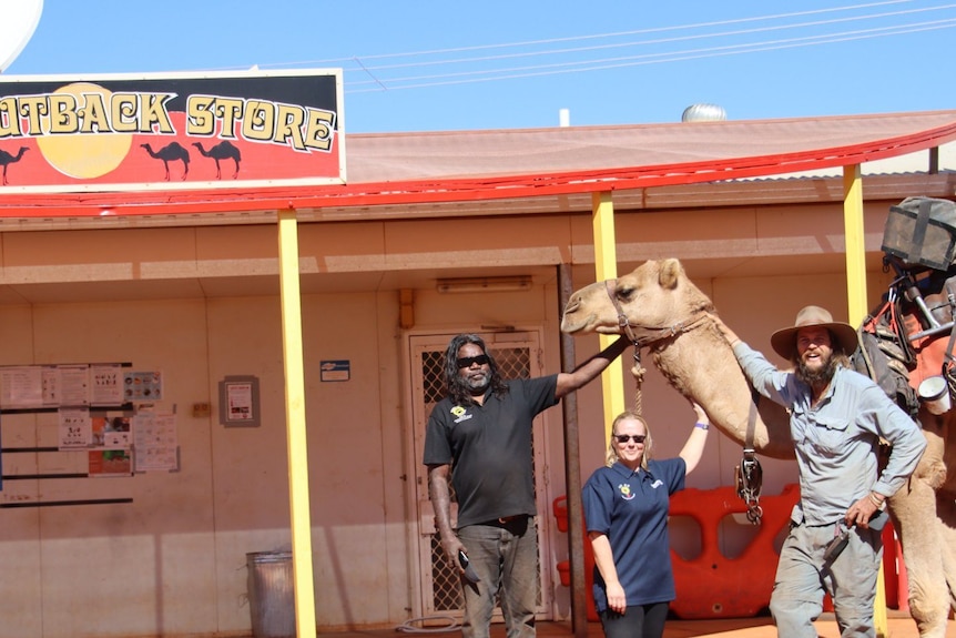Three people stand next to a camel in front of a building with the sign "JJ's Outback Store". 