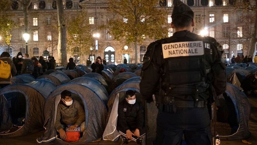 A police officer stands in front of migrants in a makeshift camp set up on Place de la Republique in Paris.