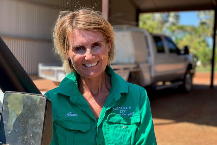 A woman in a green shirt embroidered with the name 'Jane' and 'Barkly ZTZ' leans on a truck outside an outback shed.