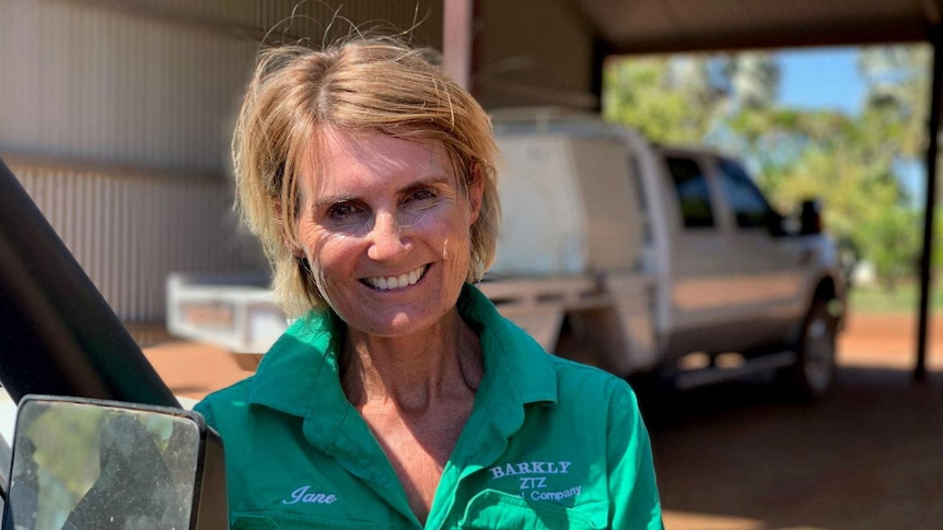 A woman in a green shirt embroidered with the name 'Jane' and 'Barkly ZTZ' leans on a truck outside an outback shed.