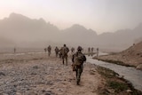 More than a dozen soldiers walking across rocky plains, with a small river running alongside and mountains in the distance.