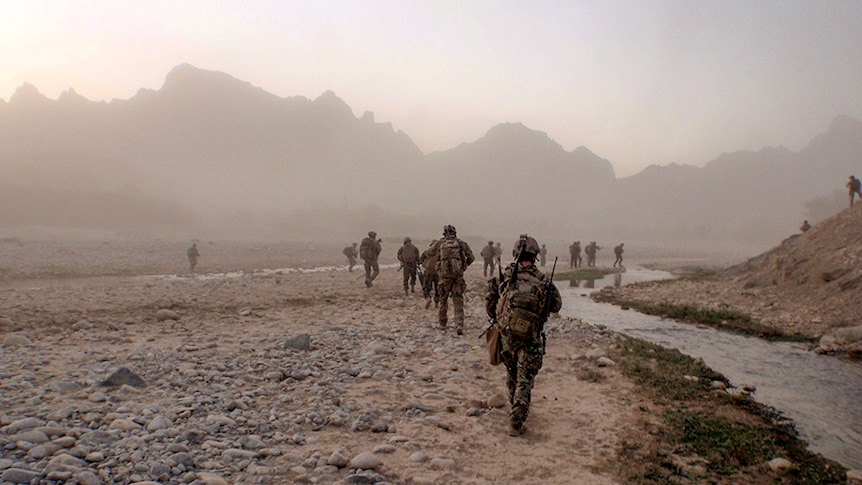 More than a dozen soldiers walking across rocky plains, with a small river running alongside and mountains in the distance.