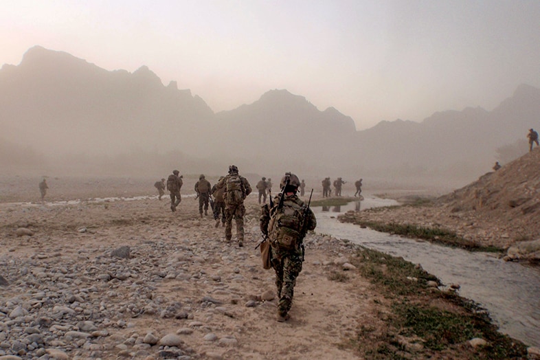 More than a dozen soldiers walking across rocky plains, with a small river running alongside and mountains in the distance.