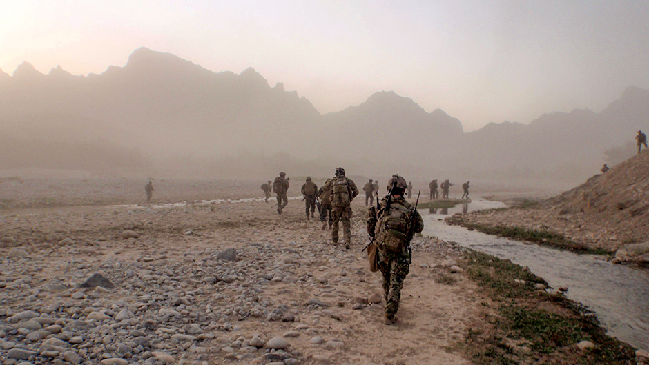 More than a dozen soldiers walking across rocky plains, with a small river running alongside and mountains in the distance.